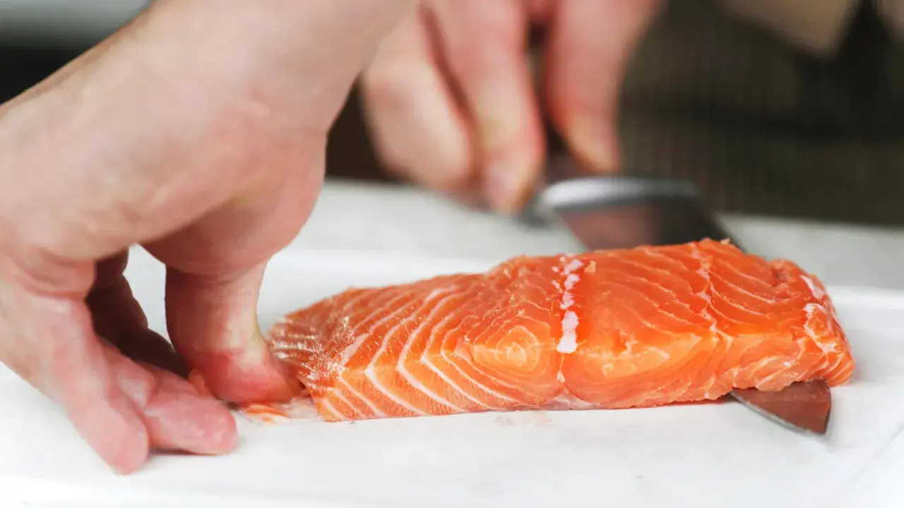 Hände halten ein Lachsfilet, während ein Messer die Haut vom Lachs entfernt.