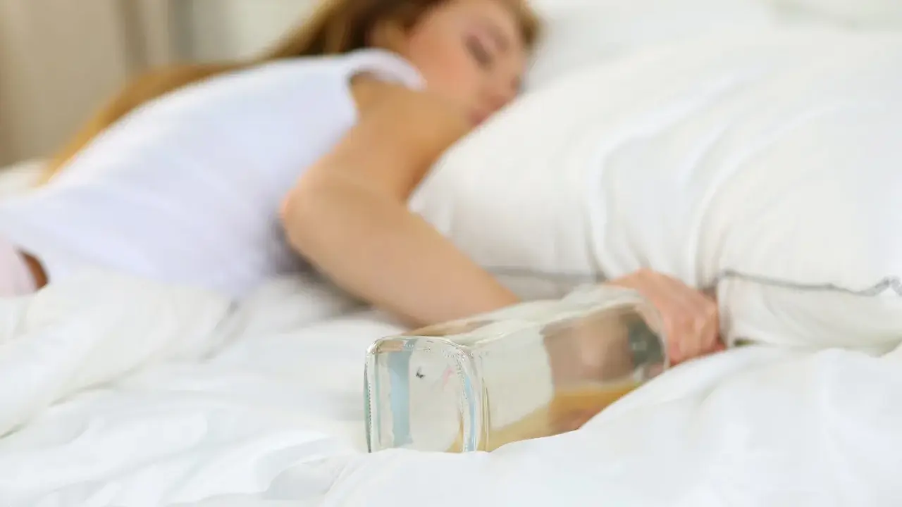 Kobieta śpi w łóżku z butelką alkoholu. Problemy ze snem mogą być związane z neuropatią alkoholową.