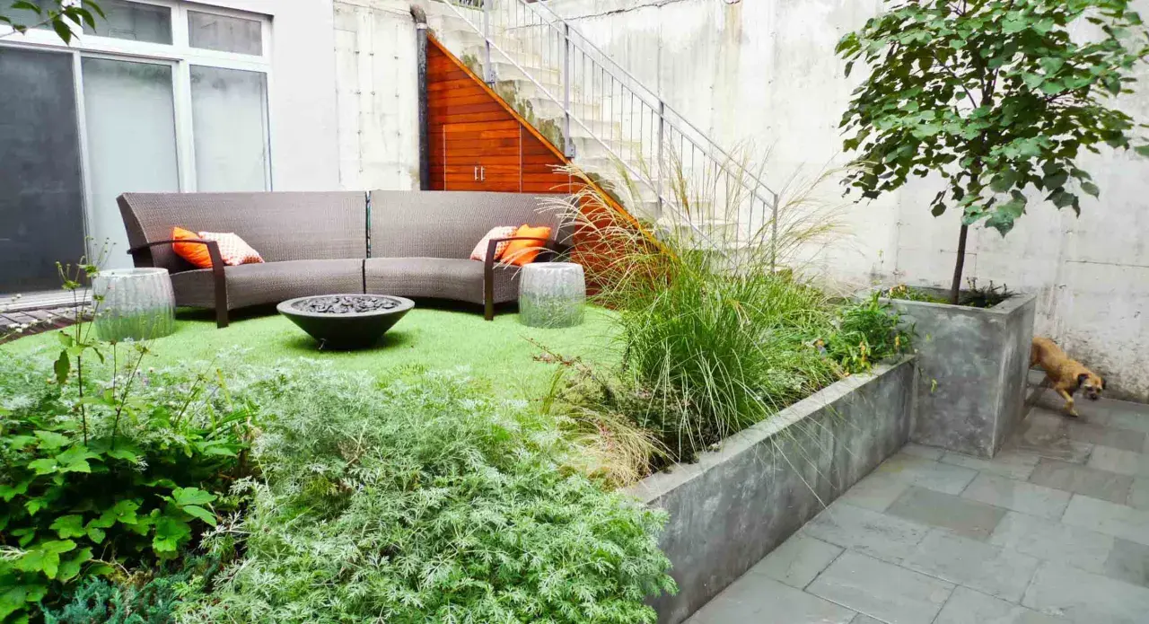 Un coin béton transformé en jardin : canapé, feu de camp, plantes luxuriantes et un chien curieux.