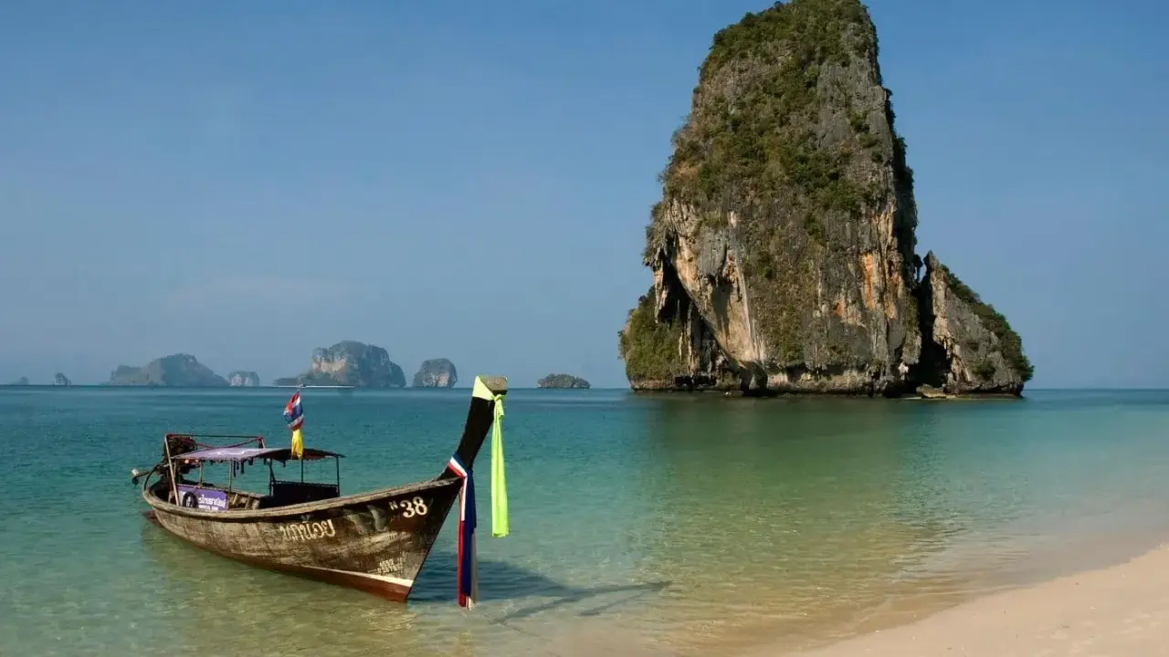 En traditionell longtailbåt guppar i det turkosa vattnet vid en strand i Thailand. I bakgrunden reser sig en grönskande klippö.