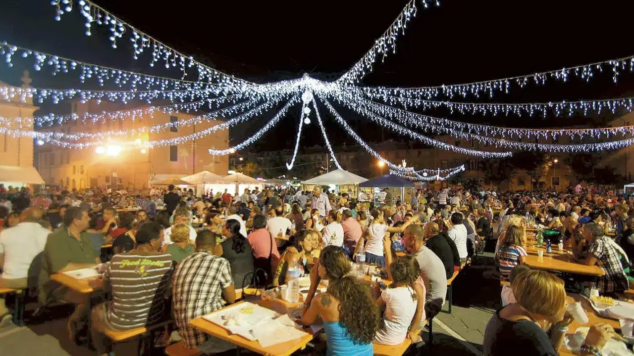 Festa notturna con migliaia di persone sedute ai tavoli, illuminate da festose luminarie. Si celebra la sagra te lu purpu, un evento conviviale.