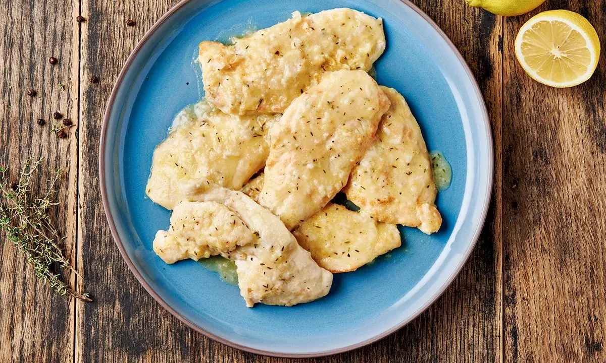 Piatto di pollo al limone su un piatto blu, con limone fresco e rosmarino sullo sfondo.