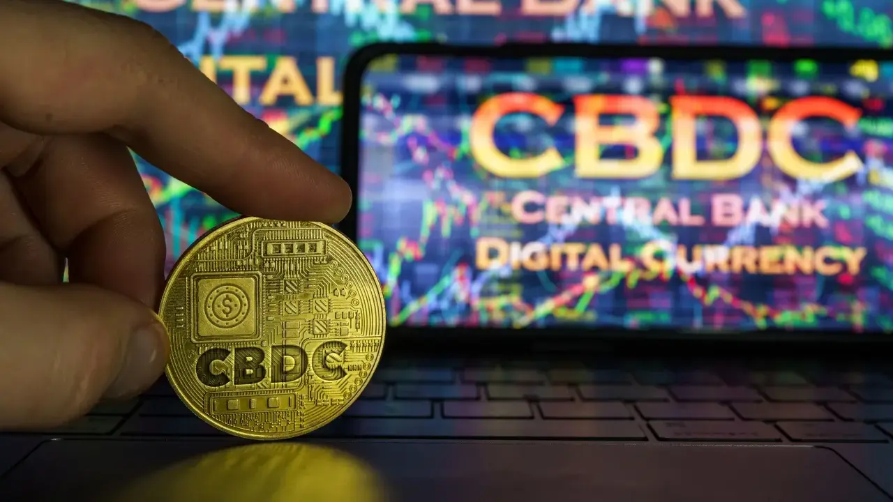 Złota moneta z napisem CBDC w dłoni, na tle ekranu z wykresem i napisem "Central Bank Digital Currency".