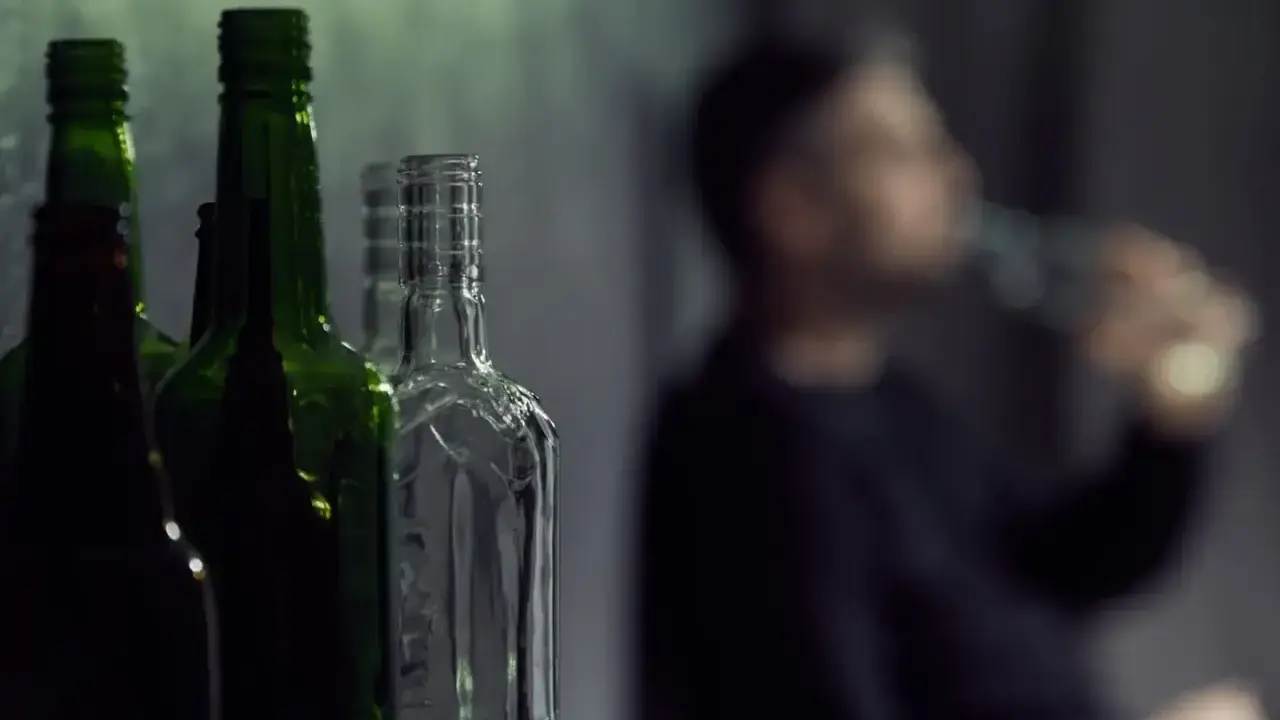 Puste butelki na pierwszym planie, w tle niewyraźna postać pijąca alkohol. Gdzie jest bezpieczna ilość alkoholu?