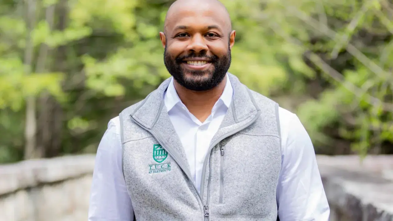 A smiling man with a beard wears a grey vest over a white shirt. The vest has a Tuck at Dartmouth logo, suggesting a curated professional image.