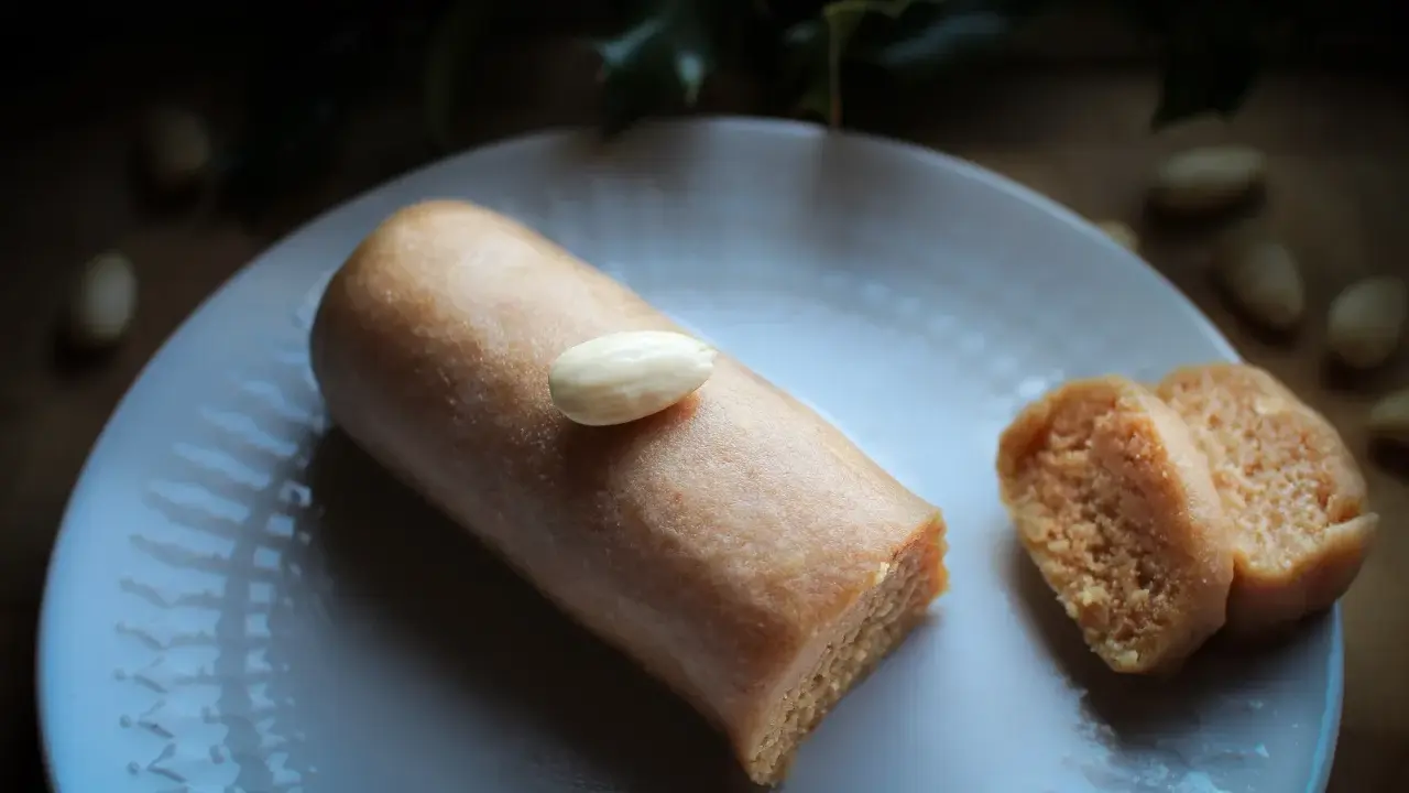 Un bâtonnet de pâte d'amande, garni d'une amande entière, repose sur une assiette blanche. À côté, deux tranches de ce délice, parfait pour un fond de tarte amande.