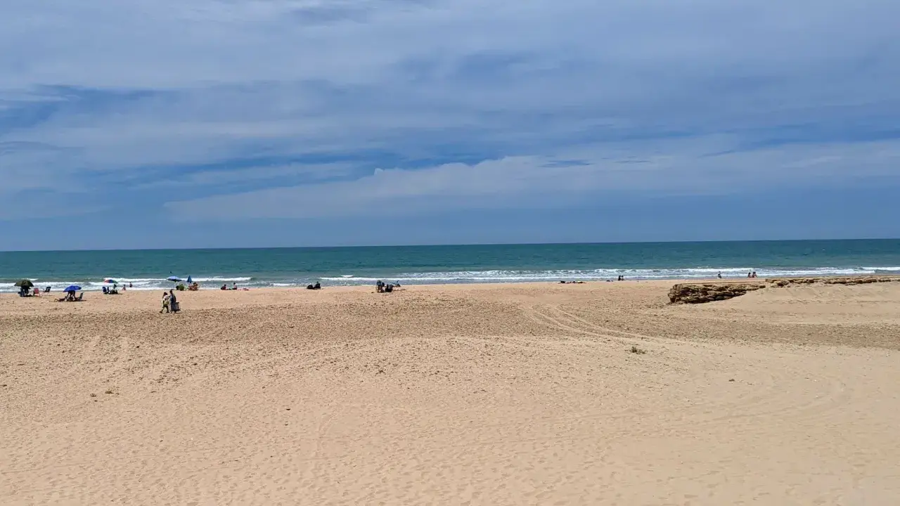 Weite Sandstrände und das Meer am Playa de Cortadura. Einige Menschen genießen den Tag unter Sonnenschirmen.
