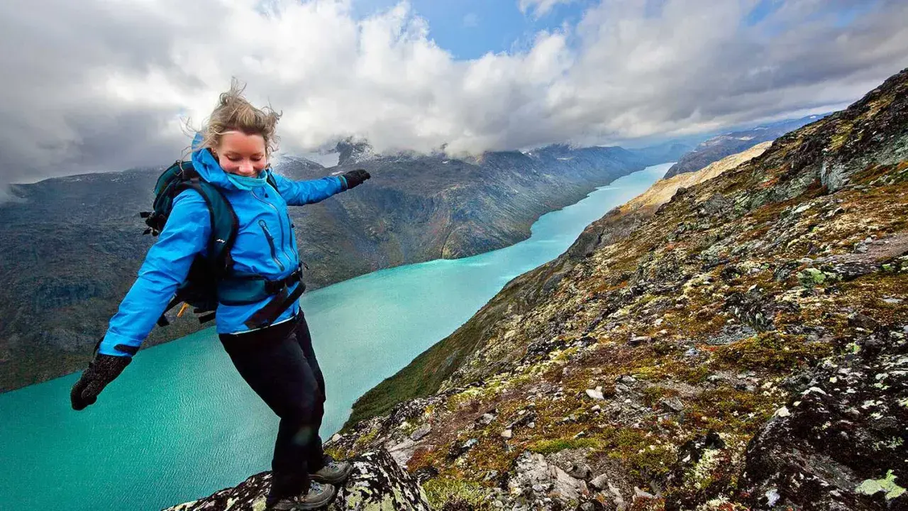 En kvinna med ryggsäck balanserar på en klippkant med en turkos sjö och berg i bakgrunden, en vy som påminner om Besseggen i Norge.