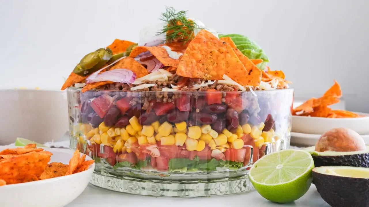 Schichtweise aufgebauter Taco Salat im Glas mit Hackfleisch, Bohnen, Mais, Tomaten, Salat, Nachos, Jalapeños und Avocado.