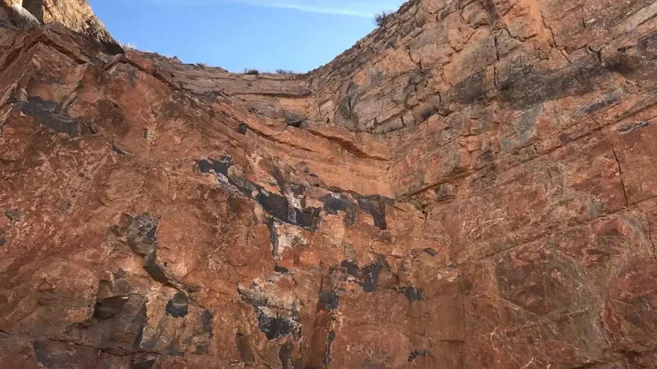 Acantilado de roca rojiza con vetas oscuras, mostrando diferentes tipos de canteras y la estratificación geológica bajo un cielo azul.