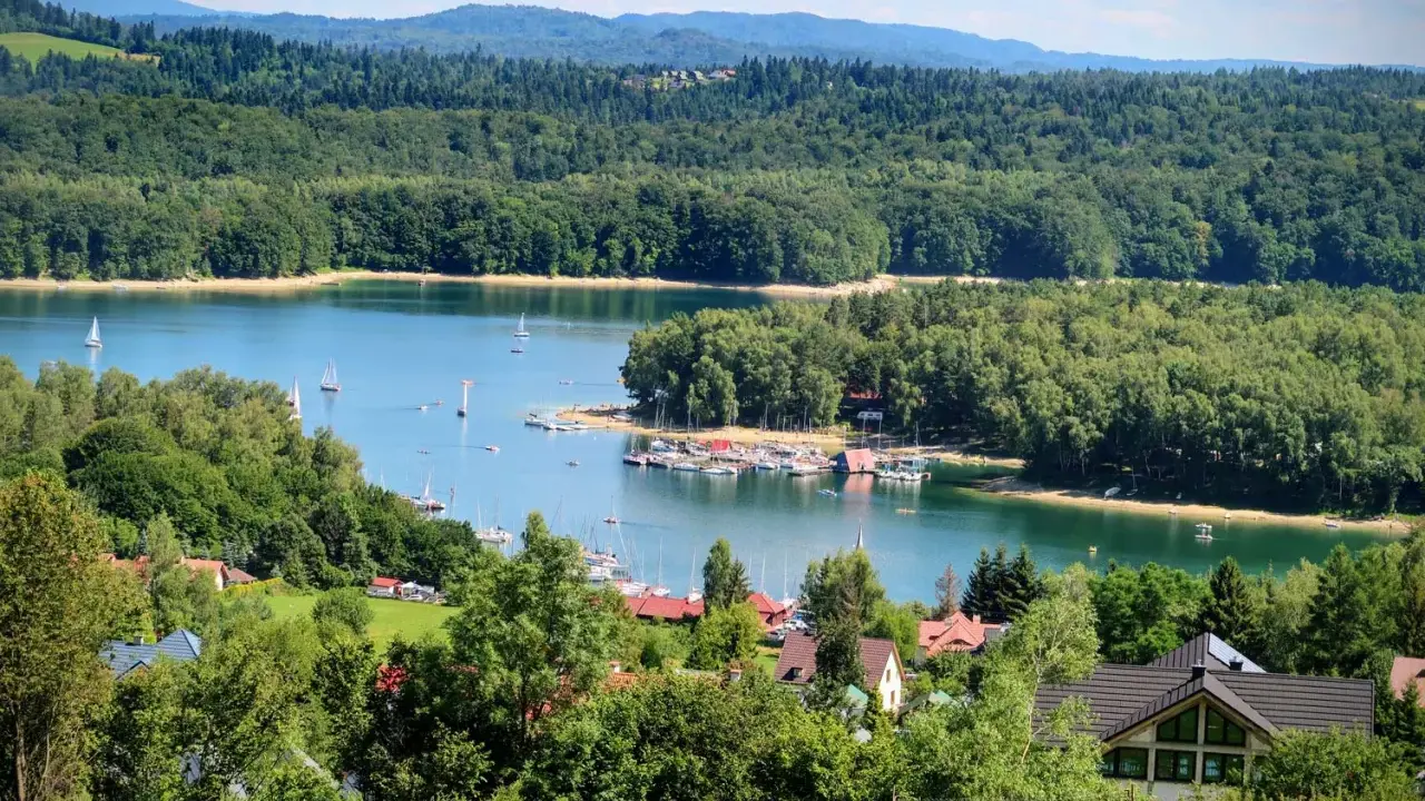 Malowniczy widok na jezioro z żaglówkami, otoczone lasami. W oddali widać góry, a na pierwszym planie domki letniskowe. Idealne miejsce na wypoczynek nad wodą, przypominające solina plaża.