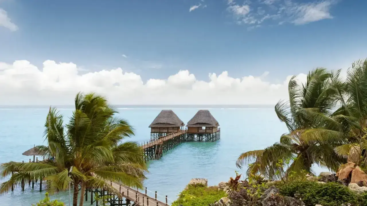 Dwa domki na wodzie połączone pomostem, otoczone palmami. Spokojne morze i błękitne niebo z chmurami. Zanzibar, pora deszczowa, ale pogoda dopisuje.