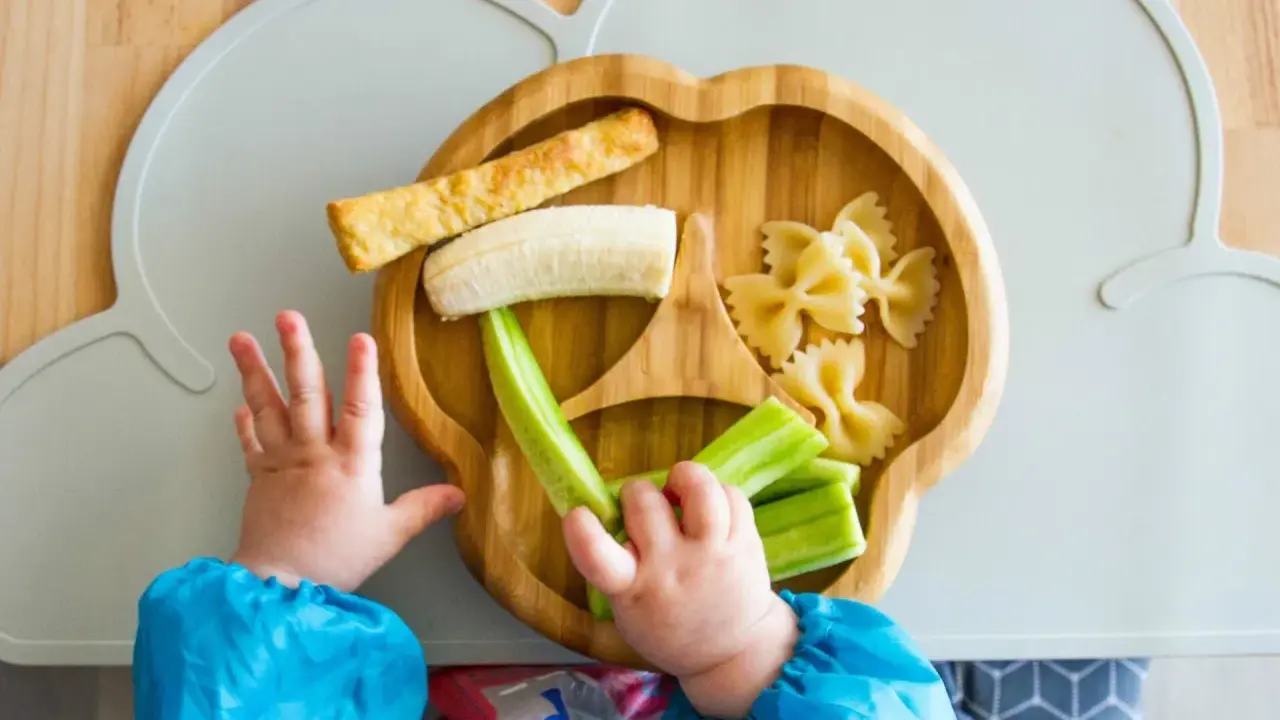 Babyhände greifen nach leckeren Baby-Snacks ab 6 Monaten: Banane, Gurke, Nudeln und ein Stück Brot auf einem Holztablett.