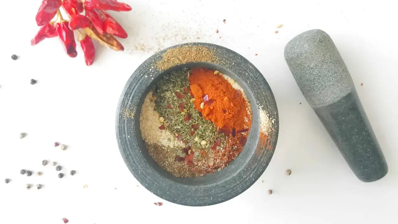 Mortier en pierre rempli d'épices pour fajitas, avec des piments rouges séchés et des grains de poivre à proximité.