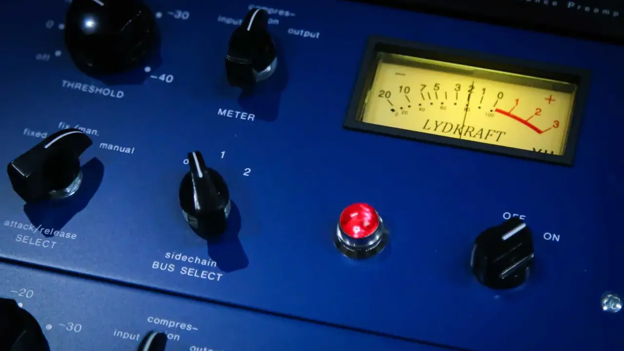 Gros plan sur un compresseur audio bleu avec des boutons, un VU-mètre et une lumière rouge allumée.