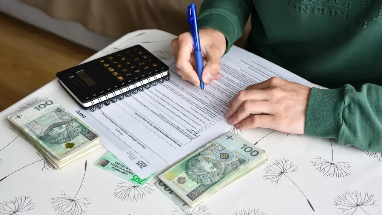 Osoba wypełnia dokumenty dotyczące umowy zlecenia i ubezpieczenia, obok leżą pieniądze i kalkulator.