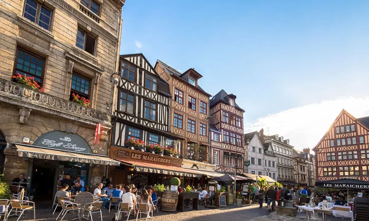 Terrasses animées devant les maisons à colombages de l'incontournable Rouen, sous un ciel bleu éclatant.