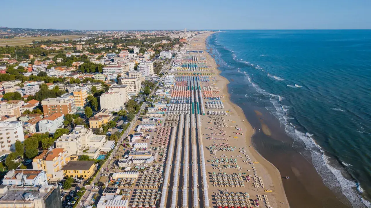 Vista aerea delle frazioni di Rimini: una lunga spiaggia attrezzata con file ordinate di ombrelloni e lettini, hotel e palazzi affacciati sul mare Adriatico.