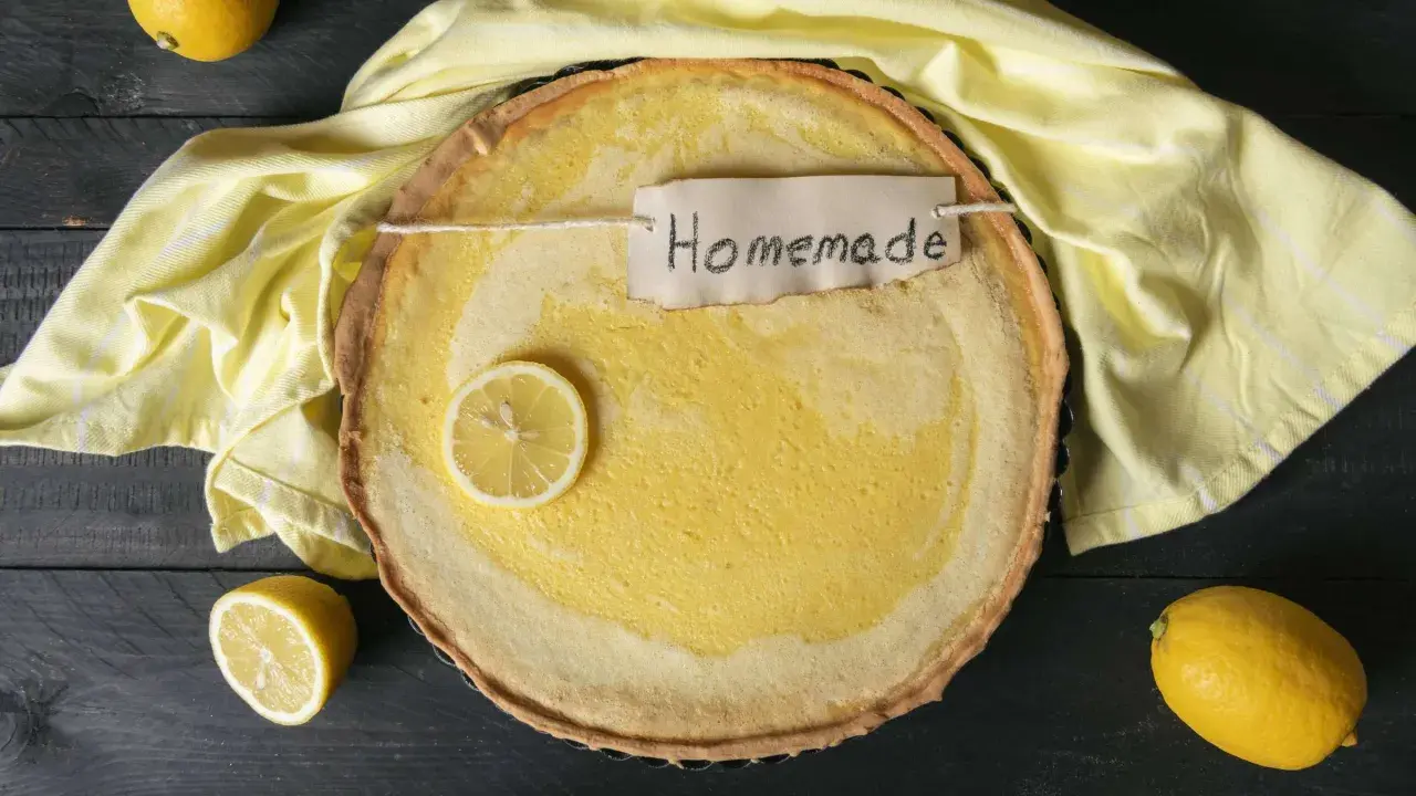 Ein hausgemachter Zitronenkuchen mit einem Zitronenschnitz und einem "Homemade"-Etikett, umgeben von gelben Tüchern und Zitronen.