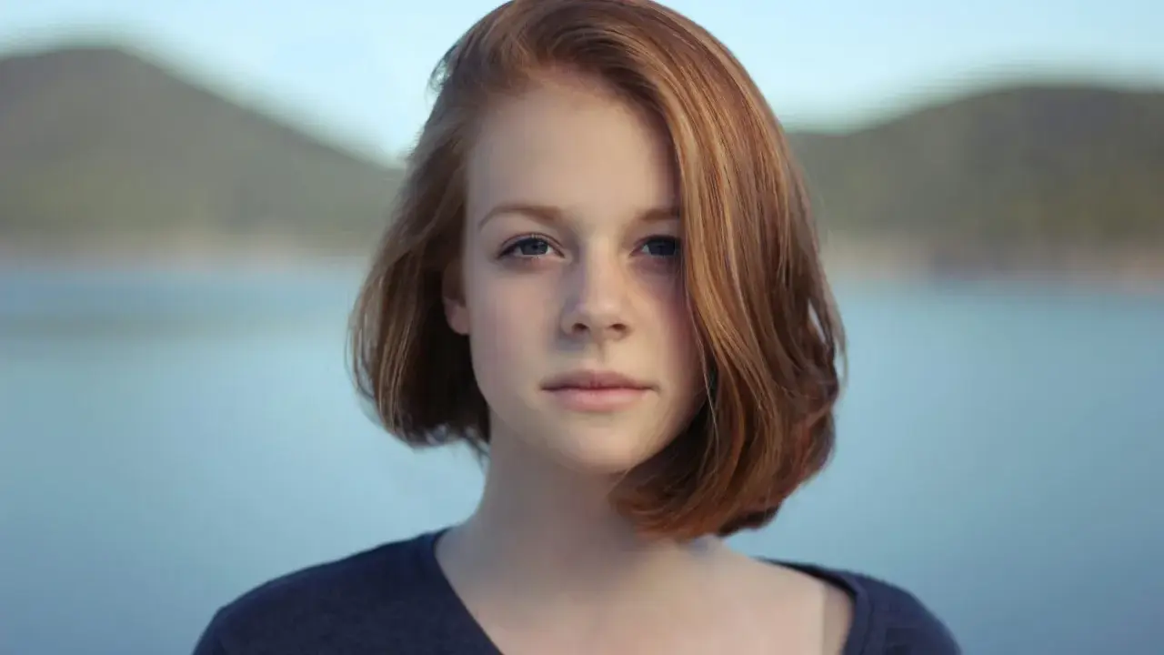 Ritratto fotografia di una giovane ragazza con capelli rossi e occhi azzurri, sullo sfondo di un lago e colline.