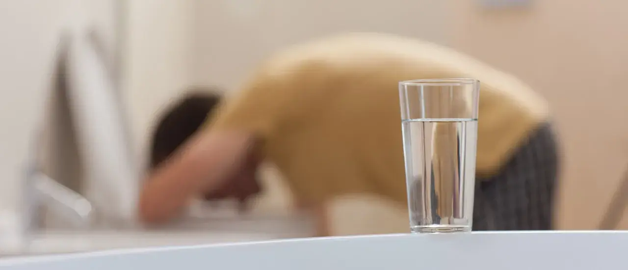 Person böjer sig över toaletten, troligen magsjuk av gammalt vatten. Ett glas vatten står på en vit yta.