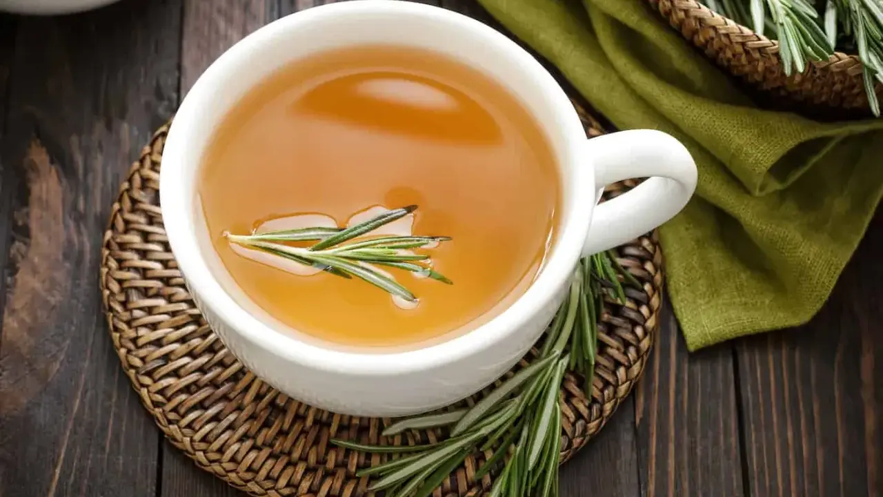 Une tasse d'infusion romarin dorée, garnie d'une branche de romarin frais, sur un dessous de plat en osier.