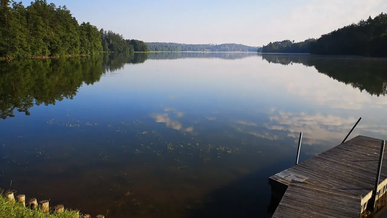 Spokojne jezioro Sulęczyno o poranku. Drewniane molo zaprasza do podziwiania odbijających się w tafli chmur i zielonych lasów.