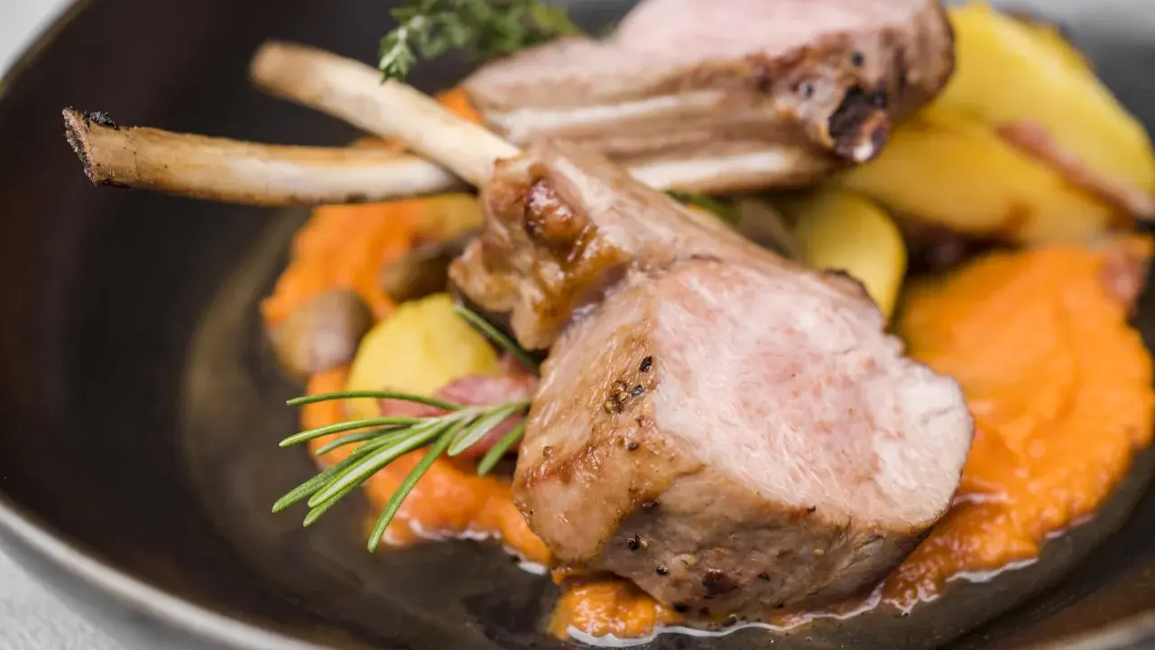 Chuleta de ternasco jugosa, servida sobre puré de zanahoria y patatas asadas, decorada con romero y tomillo.