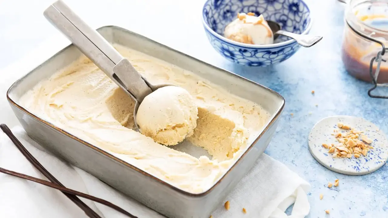 Une cuillère prélève une boule d'une glace vanille maison crémeuse dans un plat. Des gousses de vanille et des éclats de cacahuètes sont à proximité.