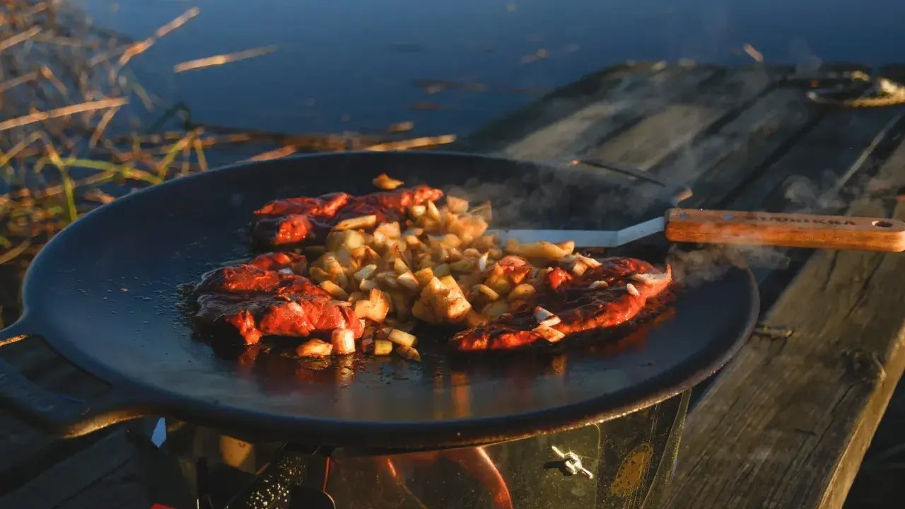 Kött och potatis steks på en rostig muurikka vid vattnet. En stekspade med trähandtag ligger bredvid.