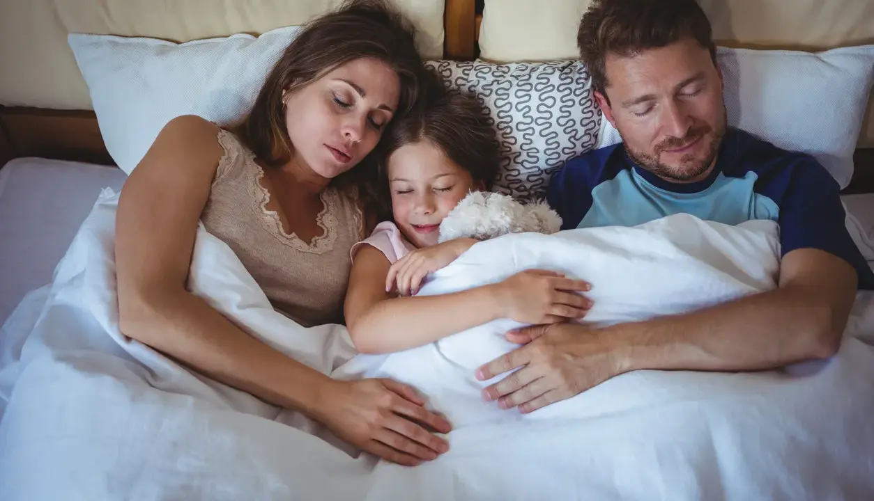 Familia durmiendo junta, la niña abraza un peluche. Un momento de paz que evoca la regresión infantil y la seguridad del hogar.