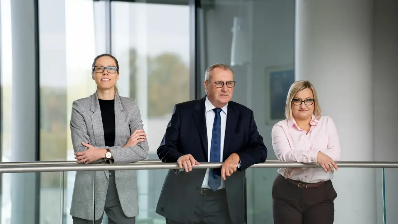 Trzy osoby w strojach biznesowych stoją przy balustradzie w nowoczesnym wnętrzu.