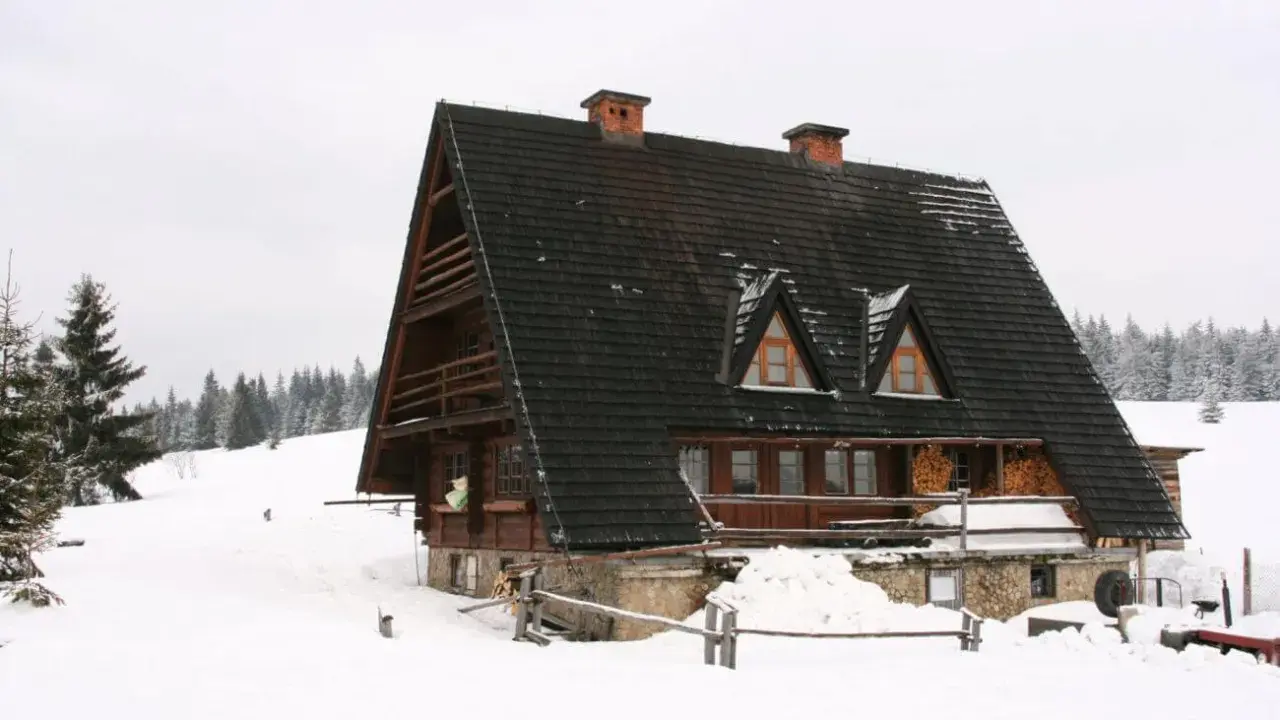 Drewniana bacówka w zimowej scenerii, otoczona śniegiem i lasem.
