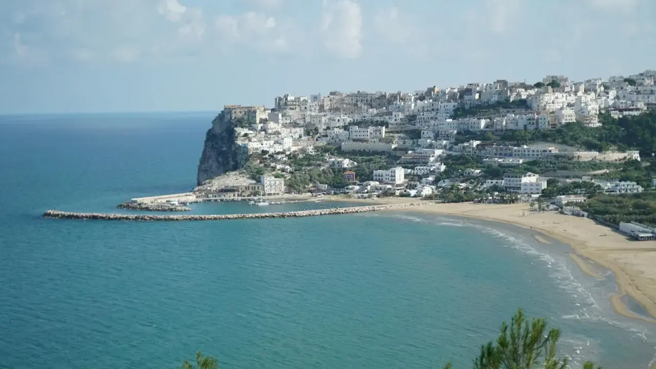 En vit stad klättrar uppför en klippa vid havet i Puglia, Italien. En strand och en hamn syns nedanför.
