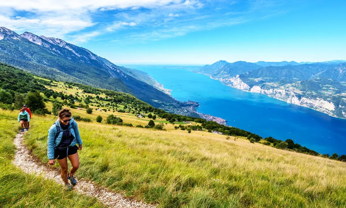Zwei Wanderer genießen die Aussicht beim Monte Baldo Wandern. Der Weg führt über einen sonnigen Hang mit Blick auf den tiefblauen Gardasee.