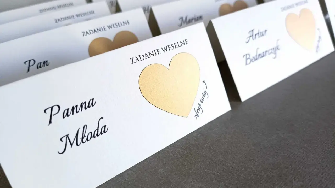 Białe karty z imionami gości i złotymi serduszkami kryją śmieszne zadania dla gości weselnych.