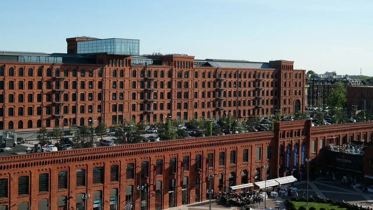 Dawna fabryka w Łodzi, dziś centrum handlowe i rozrywkowe. Widać tu architekturę z czerwonej cegły, parking, ogródki kawiarniane i karuzelę, tworząc ciekawe łódzkie atrakcje.