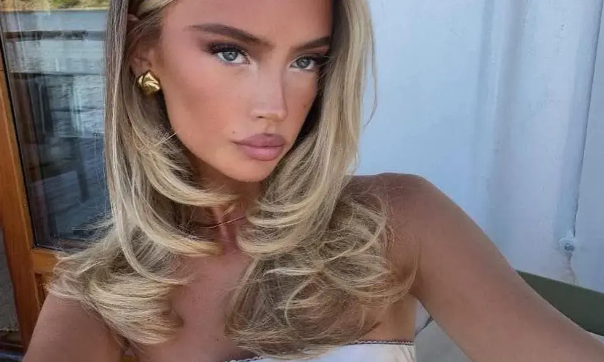 Kobieta z blond włosami, stylizowanymi na fale, z subtelnym makijażem i złotymi kolczykami. Jej fryzura przypomina buzz cut, ale jest dłuższa i bardziej objętościowa.