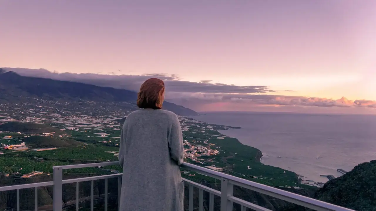 Frau blickt auf weite Landschaft, die ihr Angst vor Urlaub macht. Berge, Meer und Felder im Abendlicht.