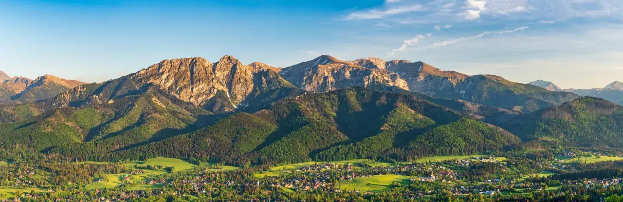 Widok na Tatry z Bachledzkiego Wierchu. Słońce oświetla skaliste szczyty i zielone zbocza, poniżej rozciąga się malownicza wieś.