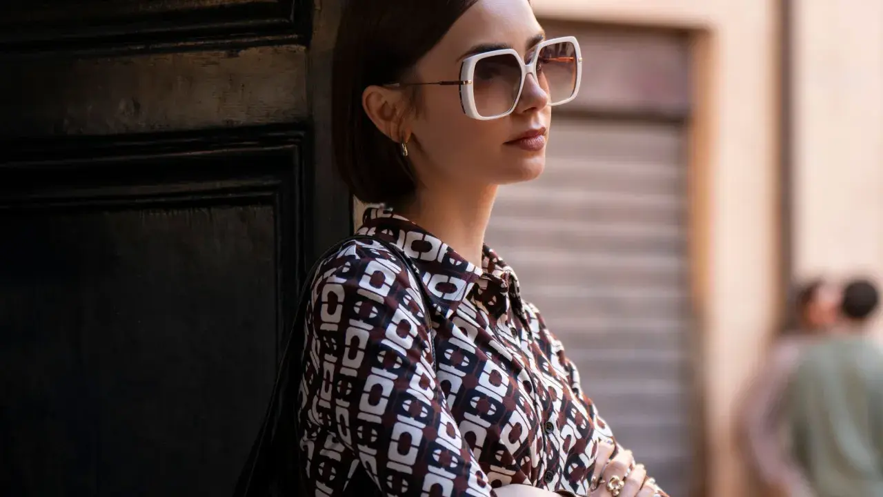 Emily in Paris, Lily Collins poses with large white sunglasses and a patterned shirt, leaning against a dark door.