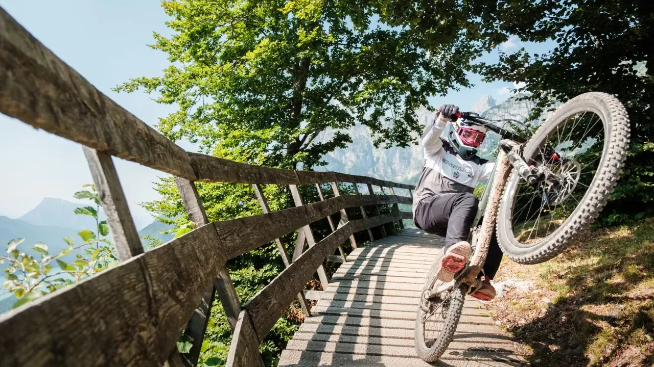 Ciclista in azione durante il giro del lago di Molveno in bici, su un ponte di legno tra alberi verdi e montagne.