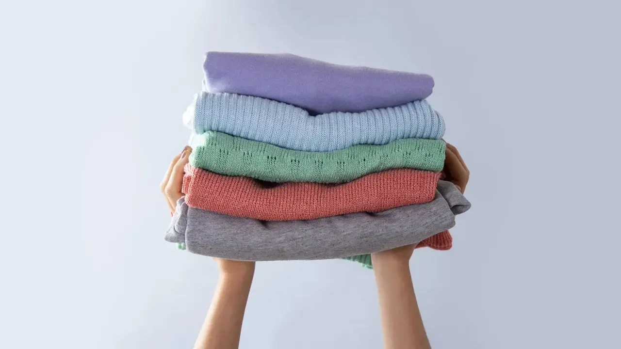 Manos sostienen una pila de ropa doblada en tonos pastel. Consejos para saber como evitar que una prenda destiña.
