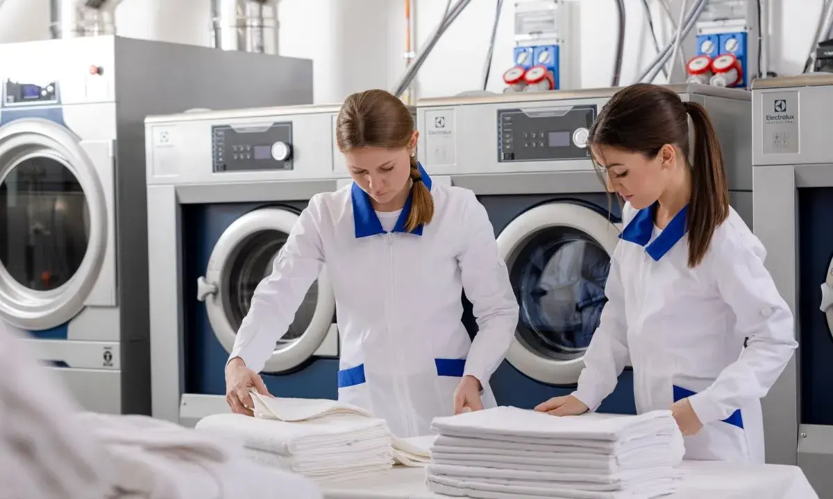 Pracownice hotelowej pralni składają czyste ręczniki. W tle widać profesjonalne pralki i suszarki.