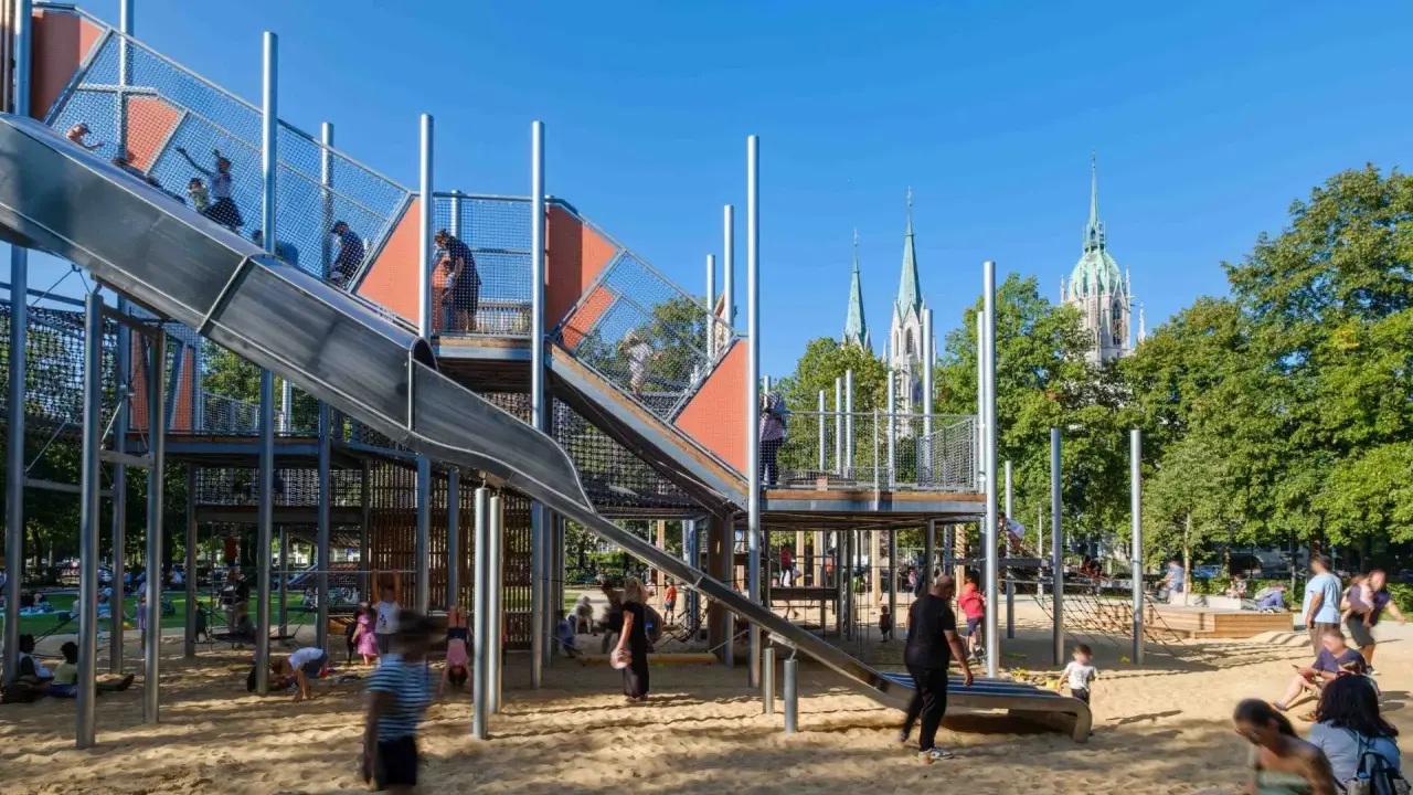 Un día perfecto de **munich con niños** en un parque infantil moderno con toboganes y estructuras de juego, con torres de iglesias al fondo.