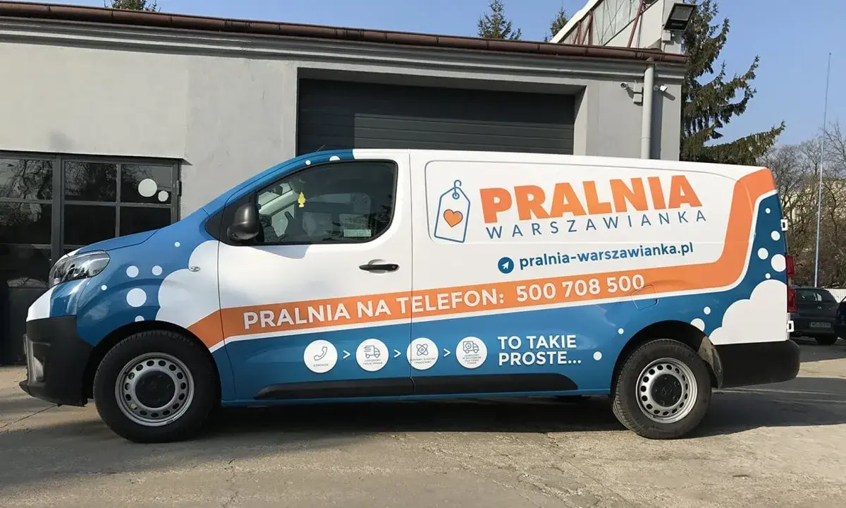 Biało-niebieski van z pomarańczowymi elementami to reklama tranzytowa dla pralni. Na boku widnieje napis "PRALNIA WARSZOWIANKA" i numer telefonu.