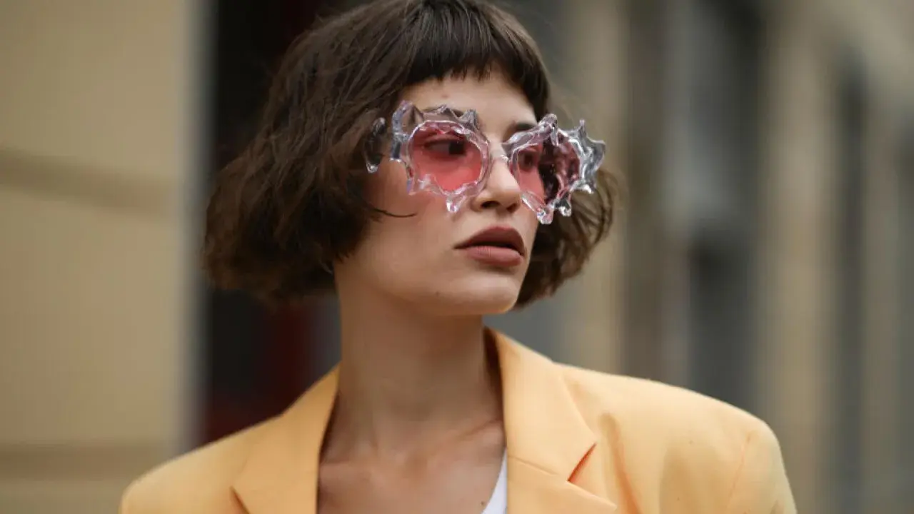 Frau mit gestuftem Bob mit Pony und auffälliger Brille mit rosa Gläsern, trägt einen hellgelben Blazer.