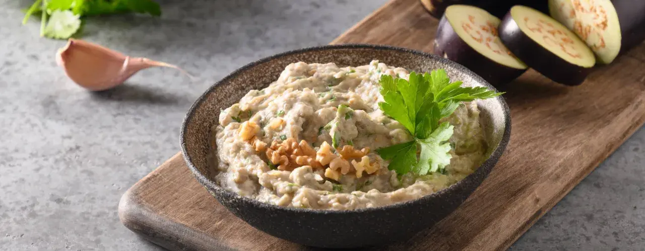 En skål med krämig baba ganoush, toppad med valnötter och persilja. Bredvid ligger skivade auberginer och ett vitlöksklyfta.