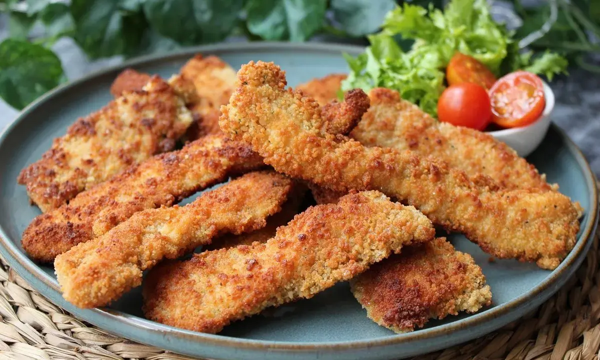 Crujientes fingers de pollo dorados en un plato azul, acompañados de lechuga y tomates cherry. ¡Perfectos para picar!