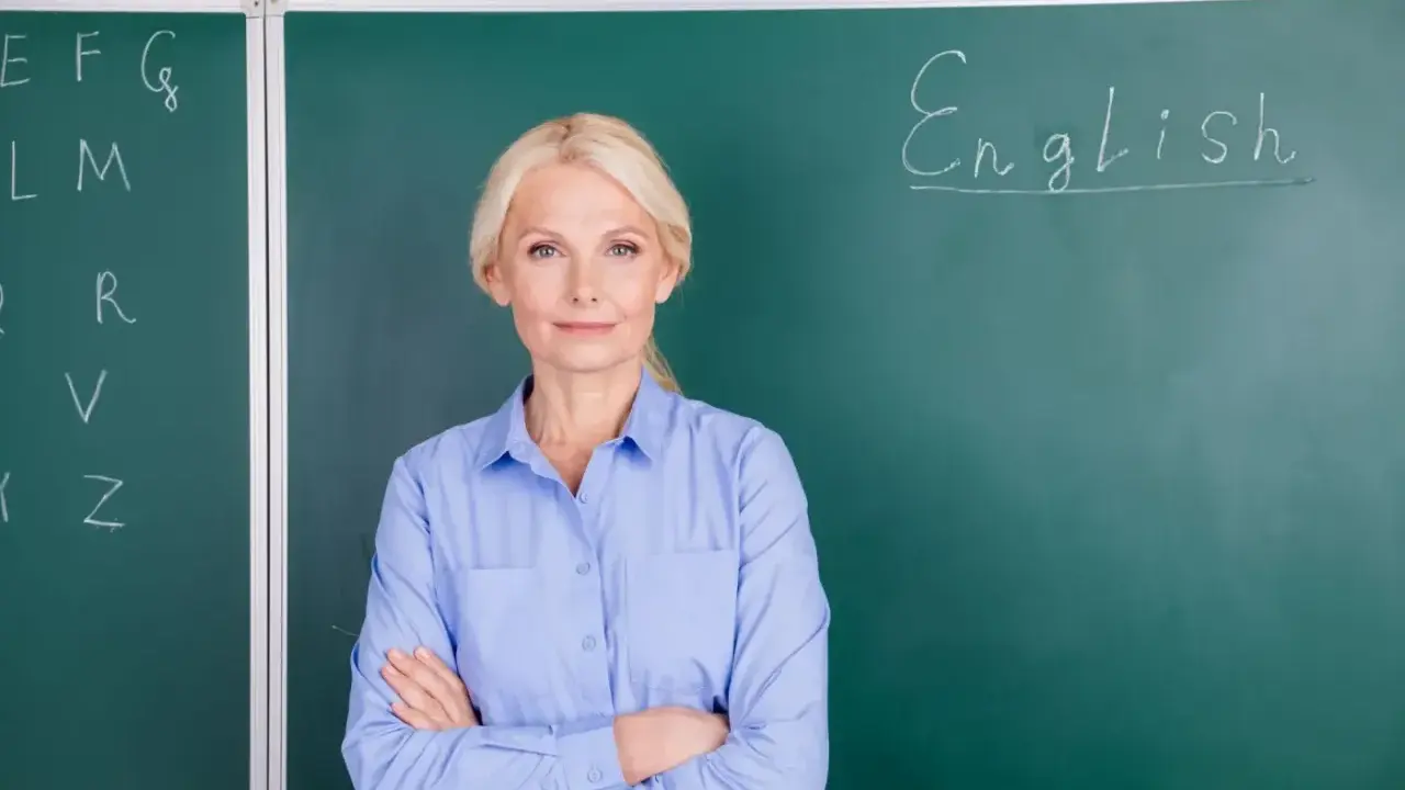 Nauczycielka angielskiego z uśmiechem, stojąca przed tablicą z napisem "English". Marzy o wcześniejszej emeryturze dla nauczycieli.